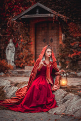 Wall Mural - Beautiful girl in a burgundy coat and red dress sitting on the background of the castle in the park, October