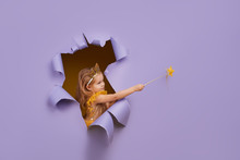 Cute Little Child Girl In Princess Costume Breaks Through A Colored Purple Paper Wall. Points With A Magic Wand To The Empty Space On The Right. Toddler Funny Emotions Face.