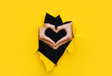 Female Hands Show A Black Heart Symbol Through The Torn Holes Of A Yellow And Black Paper Background. Creative Art, Copy Space. Concept Of Love.
