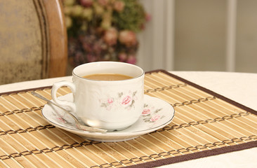 Cup with coffee on a bamboo napkin on the background of the back of a chair and a bouquet of flowers