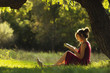 © fantom_rd - sunny portrait of a beautiful girl sitting on green glade under tree branches with book, woman reading novel on nature, concept hobby and lifestyle