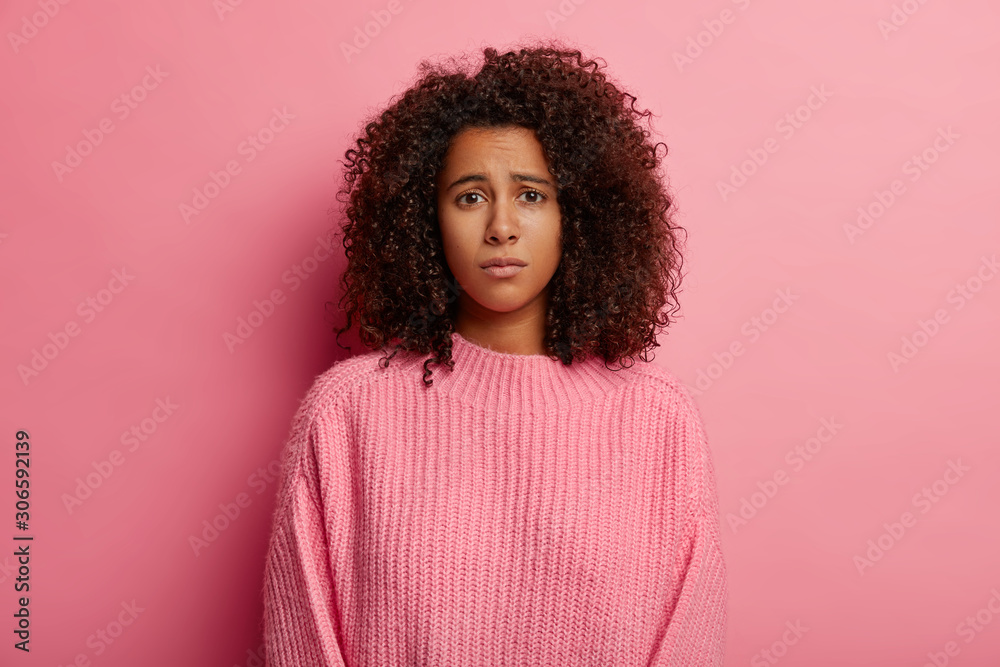 Photo of upset Afro woman has regretful look, displeased face expression, dressed in casual clothes, unhappy with bad news, looks sadly at camera, wears sweater, isolated on pink background. - obrazy, fototapety, plakaty 