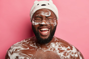 Spa, hygiene and relax concept. Naked black unshaven man takes shower with foam, has fun in bathroom, washes body, smiles from pleasure, starts day happily, enjoys morning hygiene and cleaning