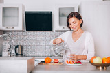 Young sincerely smiling female pouring a purified mineral water into transparent glass decanter at modern kitchen. Plenty of apples, grapefruits, kaki and oranges fruits, berries are on the tables.