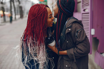 two beautiful and stylish dark-skinned girls with long hair standing in a autumn city near purple wall and drinking a coffee