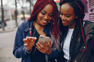 two beautiful and stylish dark-skinned girls with long hair standing in a autumn city near purple wall and using the phone