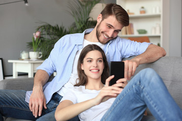 Young couple watching online content in a smart phone sitting on a sofa at home in the living room.