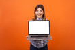 © khosrork - Portrait of glad beautiful woman with brown hair in checkered shirt standing holding laptop with blank screen and smiling at camera, place for ad. indoor studio shot isolated on orange background