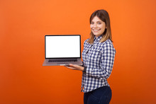 Portrait Of Happy Cute Woman With Brown Hair In Checkered Casual Shirt Standing Holding Laptop With Blank Screen And Smiling At Camera, Place For Ad. Indoor Studio Shot Isolated On Orange Background