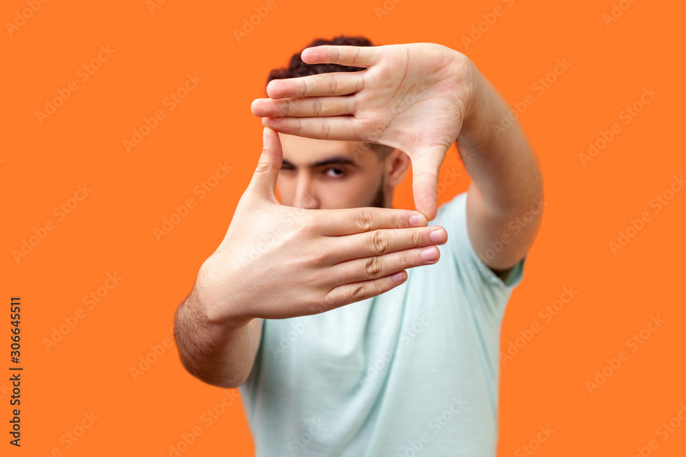 Portrait of attentive curious brunette man in casual white t-shirt looking at camera with one eye, focusing through photo frame made of fingers. indoor studio shot isolated on orange background - obrazy, fototapety, plakaty 