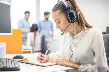 Wall Mural - Customer support operator woman talking to a client and writing notes to notebook in call center