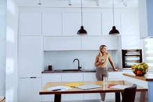 Attractive Caucasian Blonde Fashionable Woman Leaning On Kitchen Counter, Holding Mug With Coffee And Looking Trough Window.