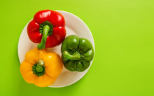 Colorful Bell Peppers On White Plate On Green Background Top View Copy Space