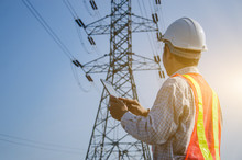 Electrical Engineer Holding And Using A Digital Tablet With High Voltage Tower Background.