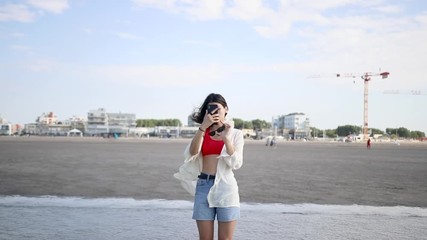 Wall Mural - young woman with denim shorts, a white shirt and a bright swimsuit walking on the beach and taking photos of the sea, ocean