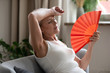© fizkes - Middle-aged woman cooling herself with hand fan indoors