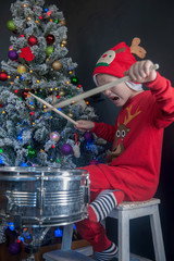 Happy Caucasian childish drummer in carnival costume Santa Claus Deer plays on a new drum set with drum sticks in his hands. Parents bought child drum as gift for Christmas. Lights on Christmas tree