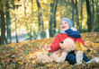 © cameravit - Little toddler boy, sitting with teddy bear in the autumn park, looking at throwing leaves around himself.