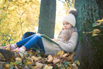 Cute little girl sitting with a book on her lap under the tree in public park. Girl sitting thoughtfully on the ground covered with yellow leaves in the autumn season sunny weather.