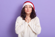 Portrait Of Sweet Attractive Young Lady Keeping Lips And Hands Together, Wearing Red Santa Claus Hat And White Sweater, Looking Directly At Camera, Standing Isolated Over Lilac Background In Studio.