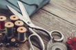 Retro sewing items: tailoring scissors, cutting knife, thimble, wooden thread spools, cushion for including pins, fabrics and sewing accessories.