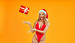 Image on isolated yellow background of young woman catch falling red Christmas gift box. Smiling excited sexy girl in red lingerie and Santa hat wearing warm gloves waiting for New Year surprise