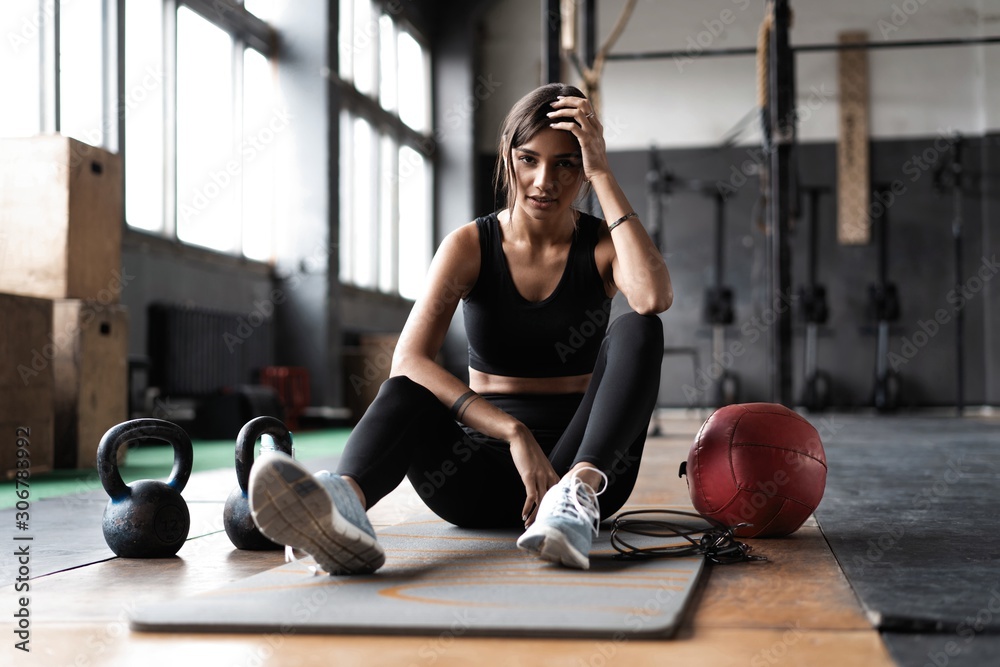 Young woman sitting on floor after her workout and looking down. Female athlete taking rest after fitness training - obrazy, fototapety, plakaty 