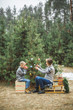 © sofiko14 - Pretty young mother and the son in gray sweaters decorating a Christmas tree in winter forest outdoors. Boy is drinking hot tea and sitting on wooden sledge with present boxes under the tree
