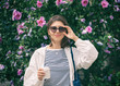 © papa - A happy young woman in sunglasses and with a paper Cup of coffee stands at a flowering Bush on the street of the summer city.