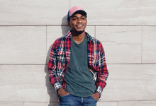 Young Smiling African Man Wearing Casuals, Red Plaid Shirt, Baseball Cap On City Street Over Gray Brick Wall Background