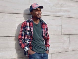 Wall Mural - Young smiling african man looking away wearing casuals, red plaid shirt, baseball cap on city street over gray brick wall background