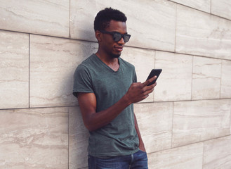 Portrait young african man with phone wearing t-shirt, sunglasses on city street over gray wall background