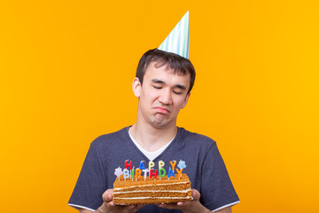 Wall Mural - Crazy cheerful young asian guy with glasses holding a burning candle in his hands and a congratulatory homemade cake on a yellow background. Birthday and anniversary celebration concept.