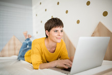 Young Woman With Laptop Lying On Bed In Bedroom Indoors At Home.