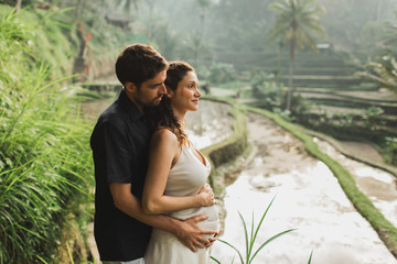 Young latin pregnant woman with husband with amazing view of Ubud rice terraces. Pregnant couple happy together.