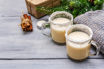 Eggnog cocktail in two glasses mug arranged with christmas decoration on light wooden table. Evergreen fir tree branch, gift, cozy jersey, artificial snow