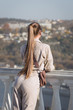 Young woman with ponytail stands leaning on balustrade and looks into distance, rear view