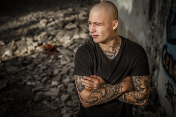 A young handsome bald brutal guy stands against the backdrop of the ruins of a building. Tattooed attractive man with arms fully covered by tattoos in a ruined building. Concept of crime, hooliganism.