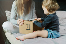 Baby And Woman Sitting On Bed And Opening A Mail Box With Products Ordered On Internet. Online Retailers Offering Wide Range Of Goods Delivered To Your Home Address. Safe And Quick Shipping Worldwide.