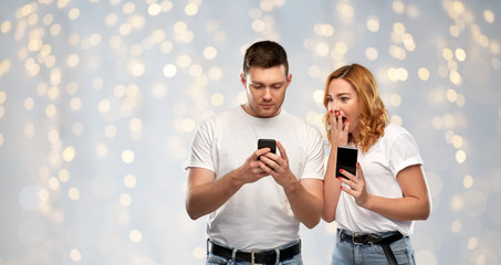 Wall Mural - technology and people concept - happy couple in white t-shirts with smartphones over holidays lights background