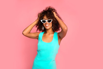 Beautiful young african american woman with afro hairstyle posing on pink pastel background wearing fashionable blue dress. Girl smiling, looking at camera with hands up. Horizontal. Copy space.