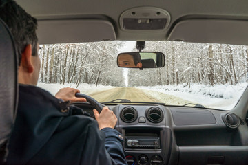 Man driving car on snowy country road through snow covered forest in winter cloudy day. Weekend roadtrip in bad weather, travel concept. Inside view, focus on landscape