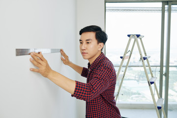 Poster - Serious young Asian man measuring walls in his new house with metal ruller before start painting