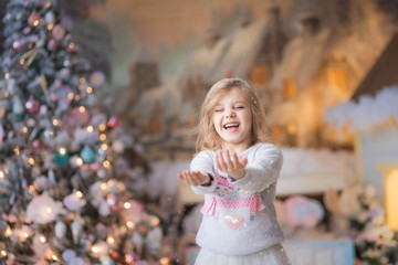 pretty happy blonde girl with curls of 6-7 years old in a thematic holiday Christmas sweater with snowmen, throws up snow and laughs, Christmas, near the New Year tree, winter