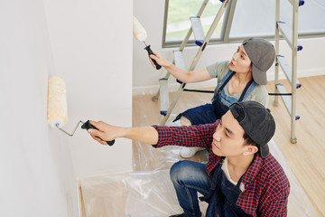 Poster - Professional young Asian wall painters in denim aprons using foam rollers when applying white paint