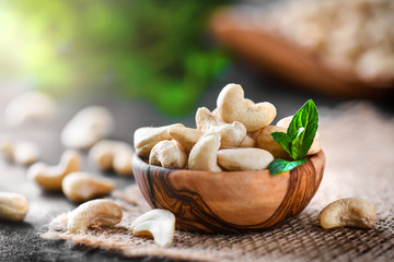 Wall Mural - Cashew nuts in wooden bowl on dark black table with mint leaf on top. Raw cashews side view.