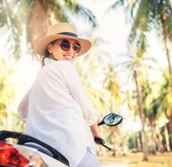 Wall Mural - Happy smiling woman in straw hat and sunglasses riding motorbike under palm tree.. Careless vacation time concept image.
