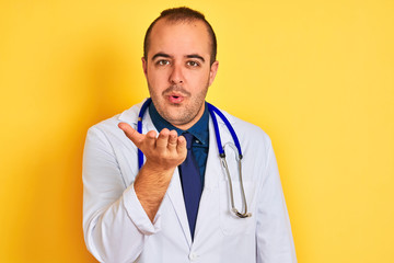 Sticker - Young doctor man wearing coat and stethoscope standing over isolated yellow background looking at the camera blowing a kiss with hand on air being lovely and sexy. Love expression.