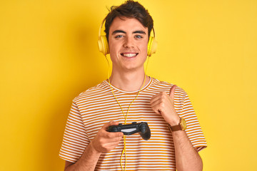 Wall Mural - Teenager boy playing video games using gamepad over isolated yellow background pointing and showing with thumb up to the side with happy face smiling