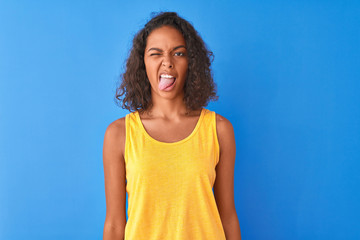 Wall Mural - Young brazilian woman wearing yellow t-shirt standing over isolated blue background sticking tongue out happy with funny expression. Emotion concept.